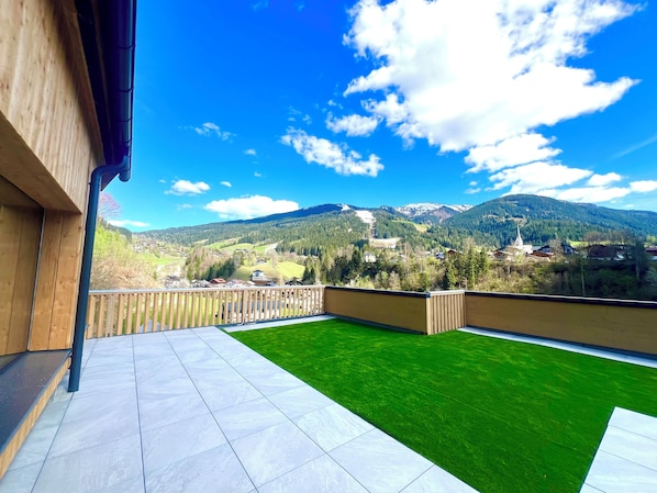 Un balcón con vistas a un paisaje montañoso, una zona ajardinada y barandilla de madera.
