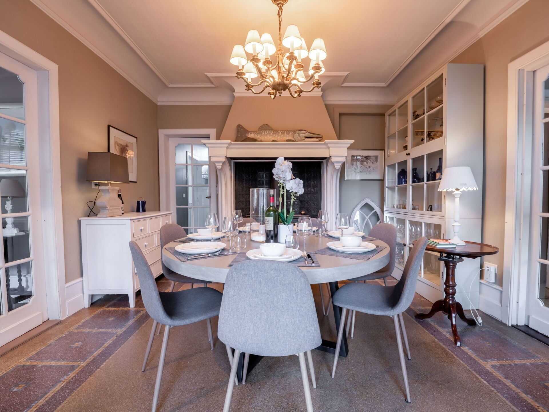 Comedor con una mesa redonda dispuesta para cuatro personas, rodeada de sillas grises. Un candelabro cuelga del techo, y hay libros y una lámpara en una mesita auxiliar.