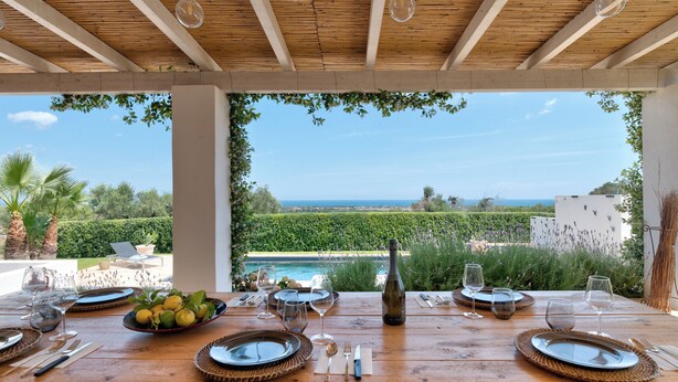Een terras met uitzicht op zee, een houten tafel gedekt met borden, glazen en een fles.