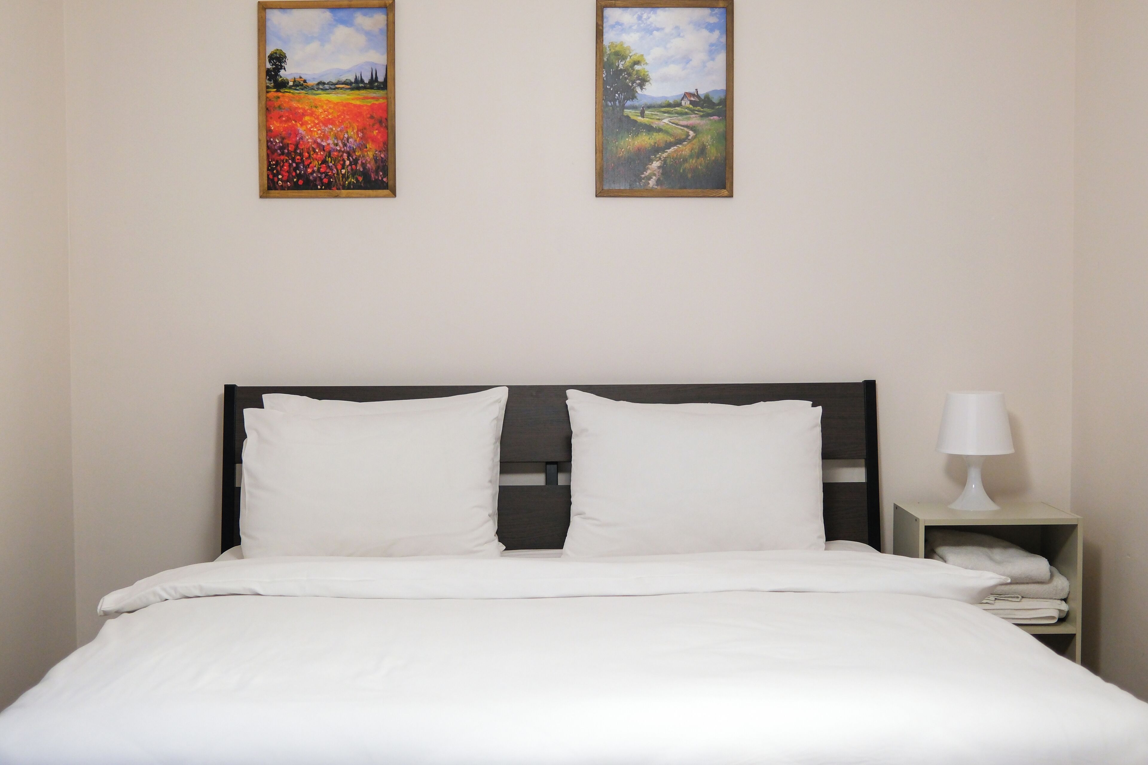 A neatly made bed with white linens, a wooden headboard, and two framed paintings on the wall above.