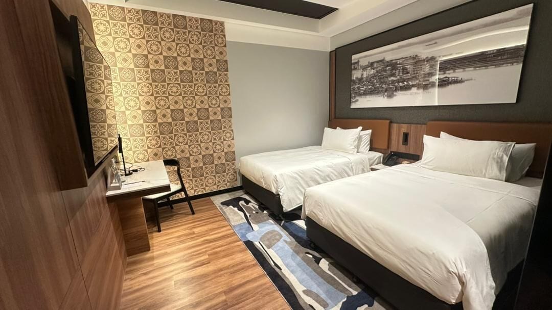 A hotel room with two beds, a desk, a chair, and a patterned wall.