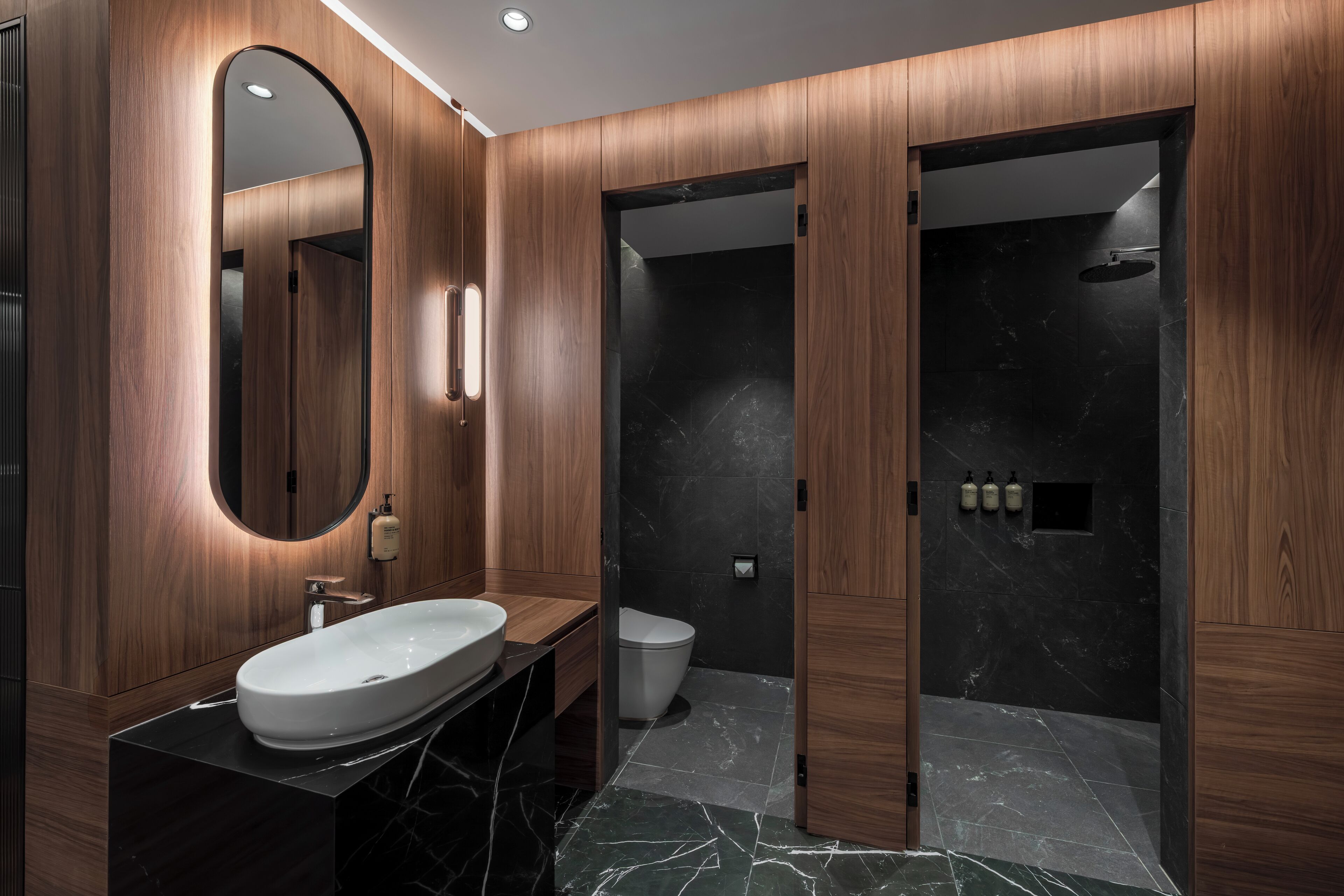 A modern bathroom with a wooden vanity, a large oval mirror, a black marble sink, a toilet, and a shower area.