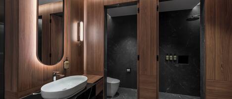 A modern bathroom with a wooden vanity, a large oval mirror, a black marble sink, a toilet, and a shower area.