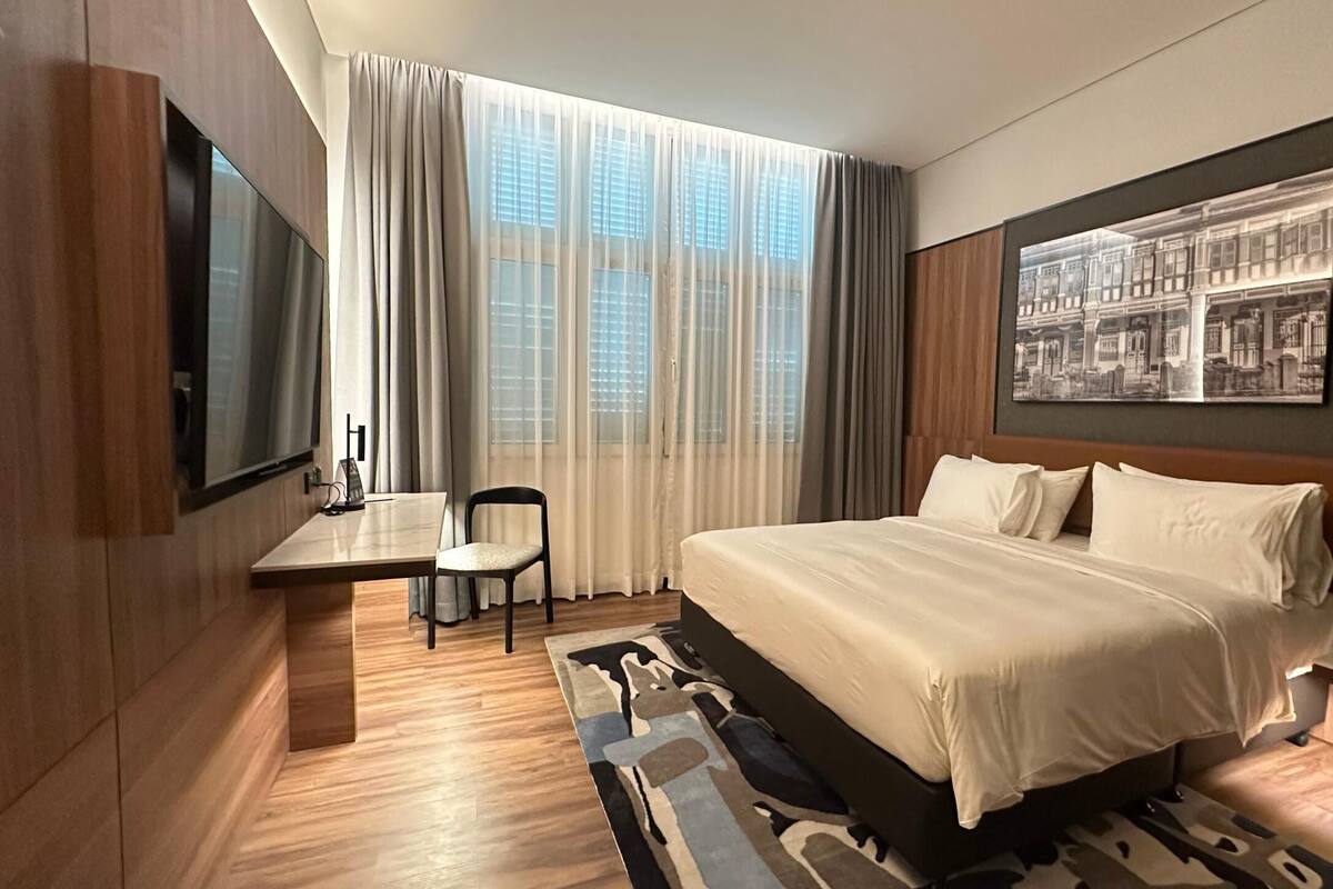 Ein Hotelzimmer mit Bett, Schreibtisch, Stuhl, Fernseher und einem großen Fenster mit Vorhängen.