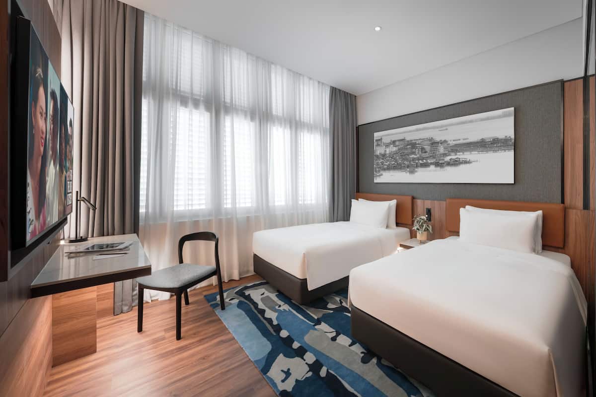 Ein Hotelzimmer mit zwei Betten, einem Schreibtisch, einem Sessel, einem Fernseher und einem großen Fenster mit Vorhängen.