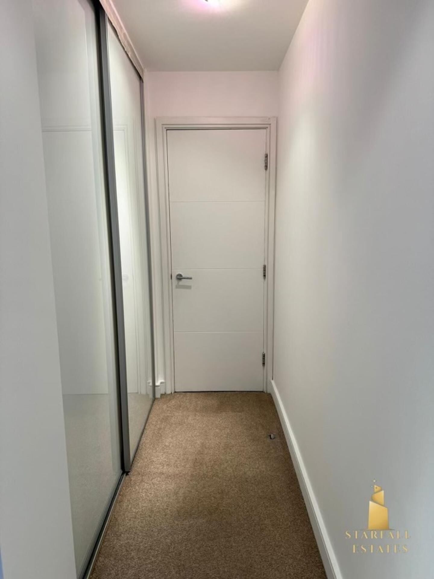Un corridoio stretto con una porta chiusa in fondo, pavimento rivestito di moquette e pareti bianche.