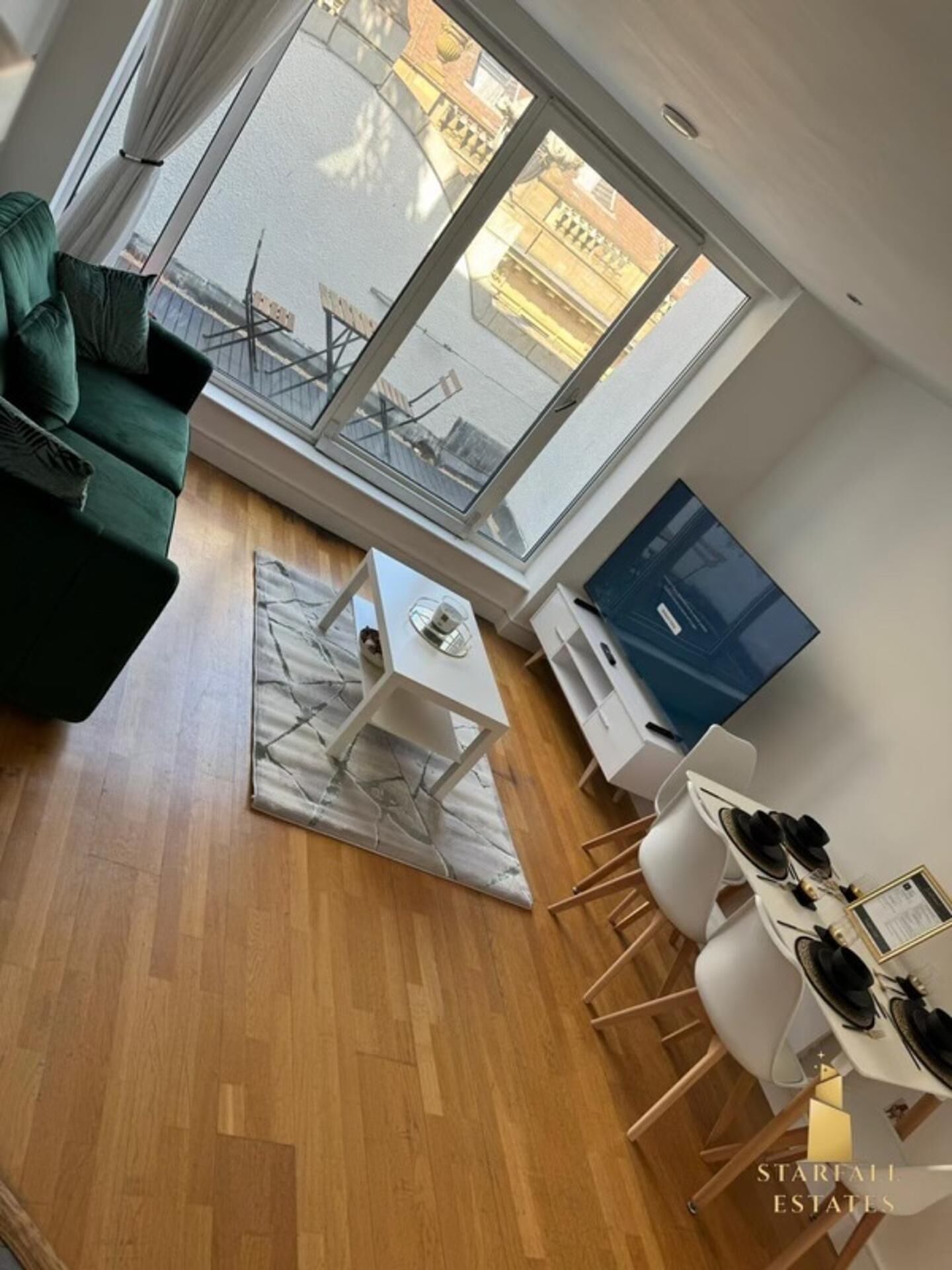 A modern living room with a green sofa, a white coffee table, and a dining area with a white table and chairs. Large windows offer a view of the street.