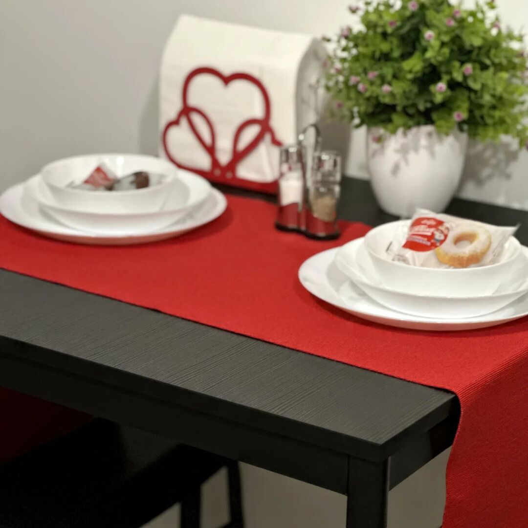 Une table dressée avec de l’assiette blanche, un chemin de table rouge et une plante en pot ornée de fleurs roses.
