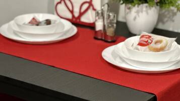 Une table dressée avec de l’assiette blanche, un chemin de table rouge et une plante en pot ornée de fleurs roses.
