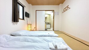 A hotel room with two beds, a television, a bedside lamp, and a small kitchenette visible through an open door.