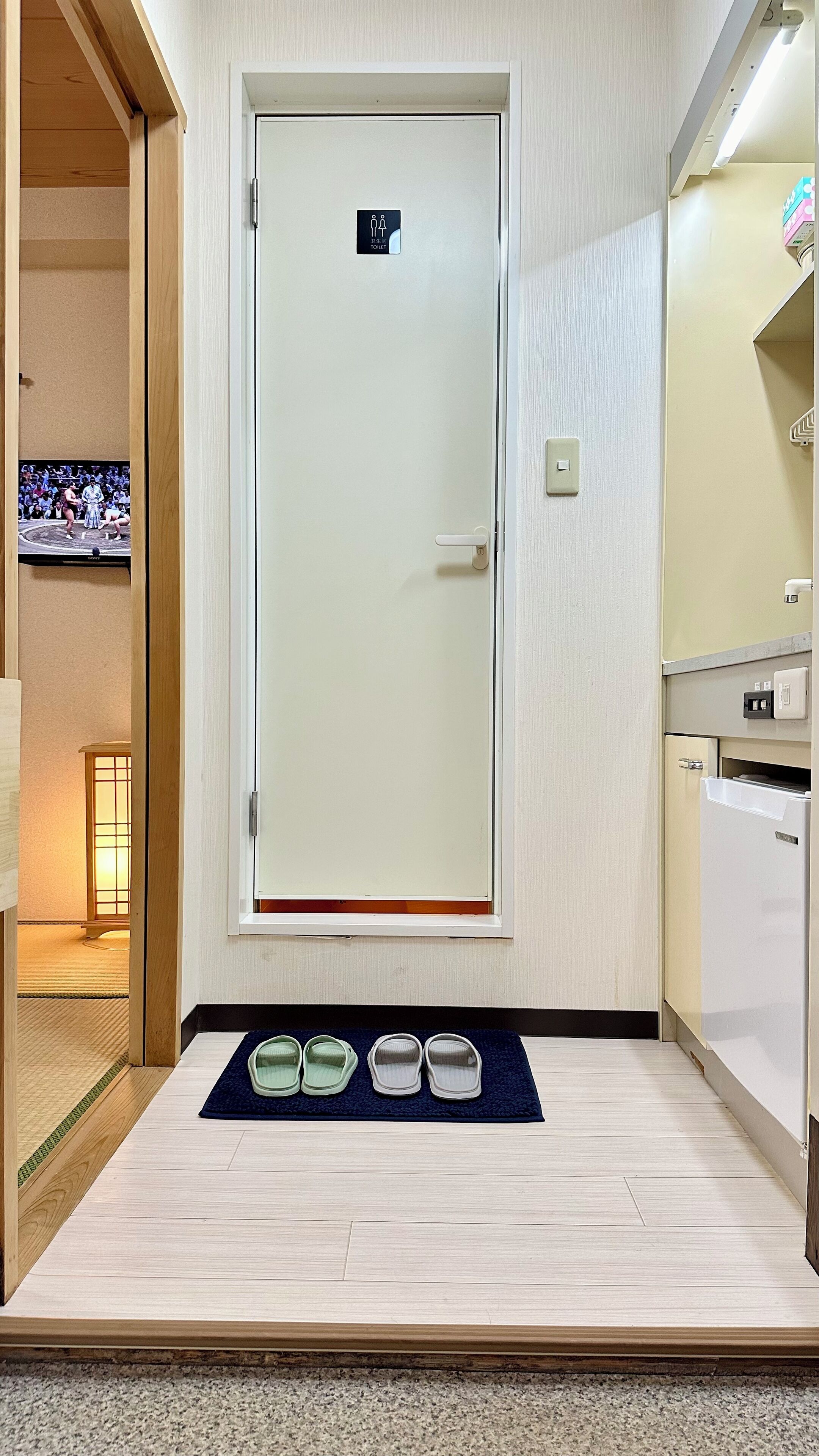 A hotel room with a wooden floor, a TV mounted on the wall, a door with a toilet sign, and a small table with slippers.
