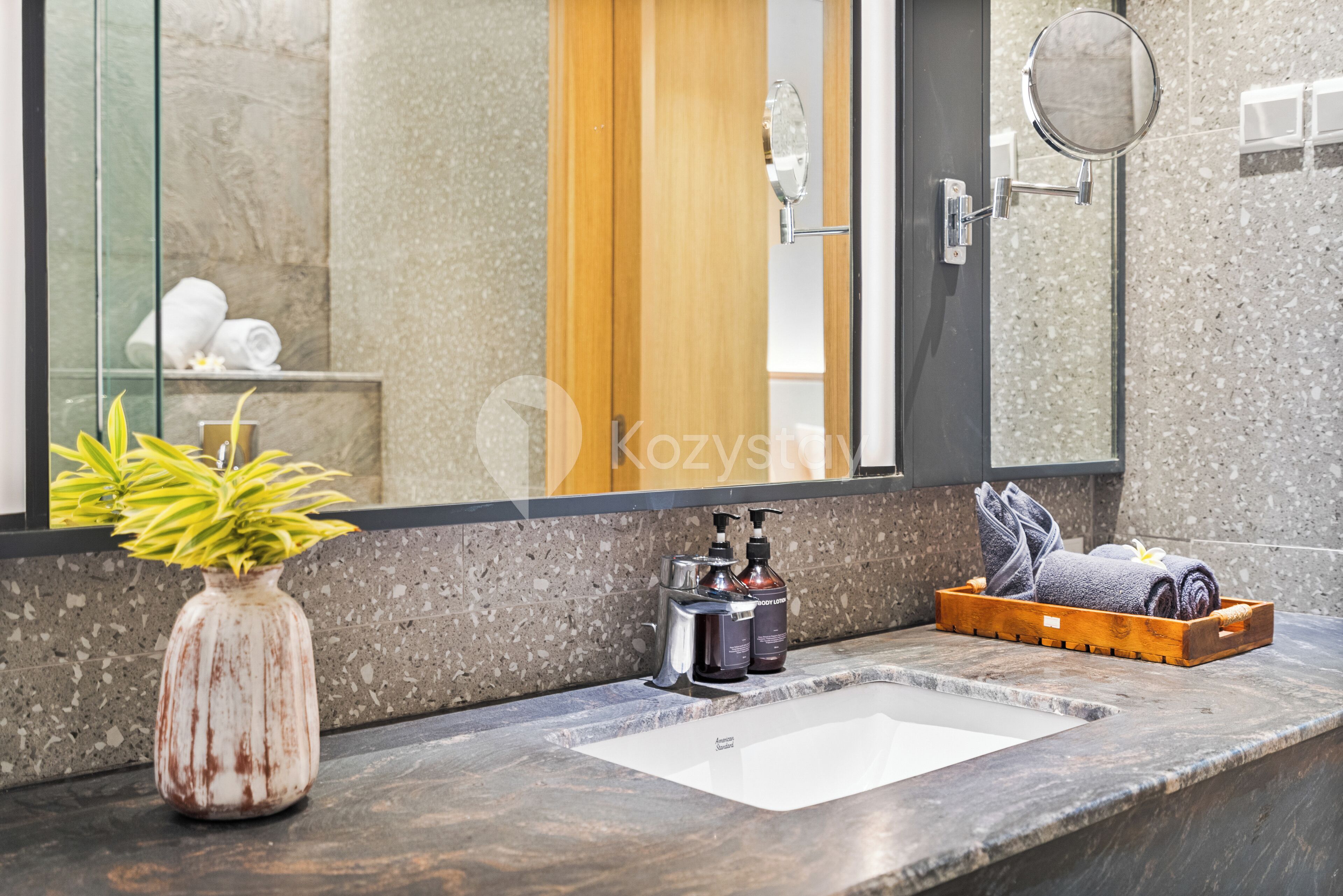 Un baño con un lavamanos de mármol, una bandeja de madera con toallas, un jarrón con una planta y un espejo.
