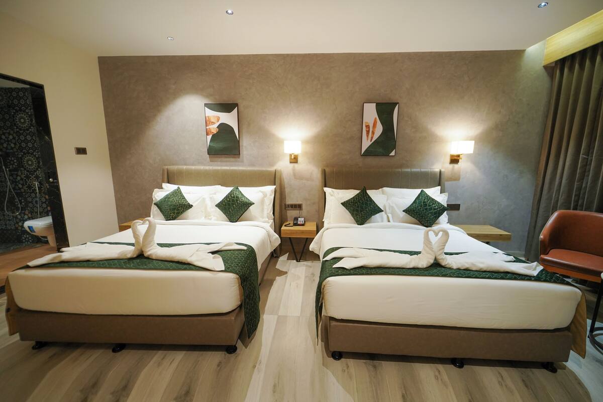 A hotel room with two beds, each with a green and white bedspread, pillows, and a heart-shaped towel arrangement. There are bedside lamps, a wooden nightstand, and a chair.