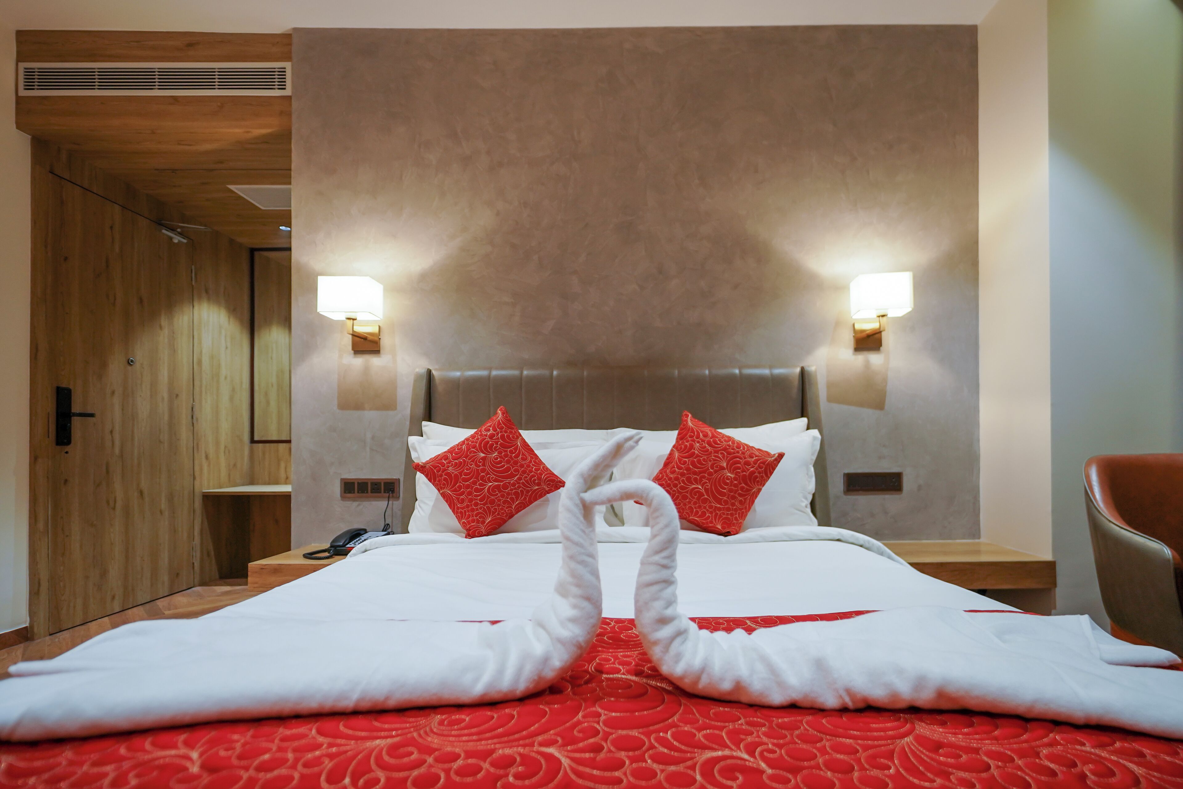 Une chambre d’hôtel avec un lit, deux oreillers rouges, une couvre-lit rouge et blanche, une porte en bois et deux lampes fixées au mur.