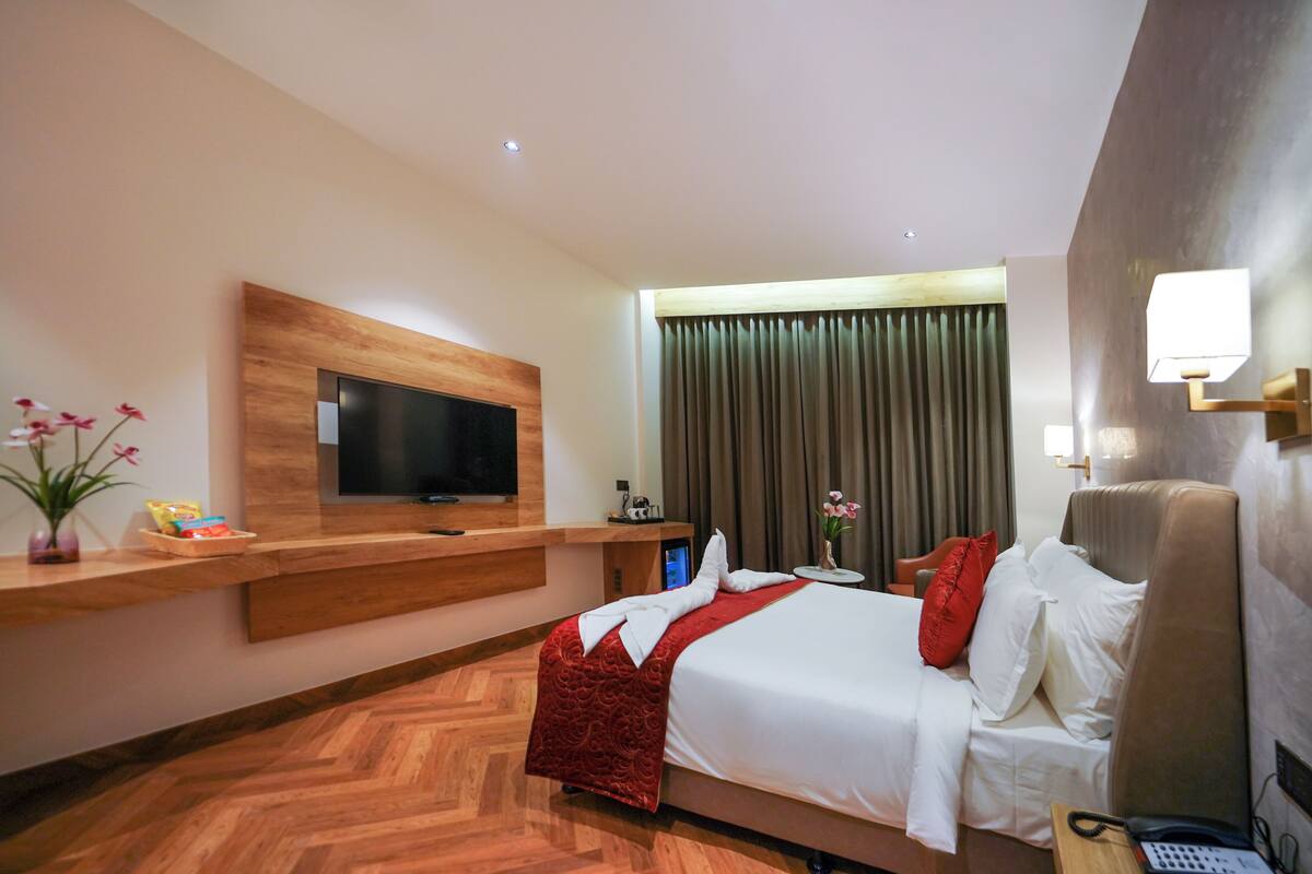 A modern hotel room with a large bed, a flat-screen TV mounted on a wooden panel, a bedside table with a lamp, and a view of the city through large windows.