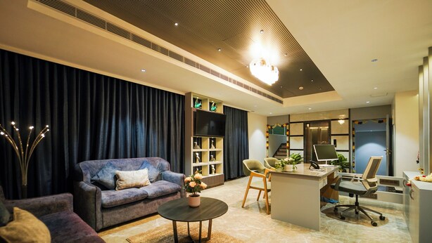 A modern living room with a sofa, a coffee table, and a desk with a computer. There is a bookshelf and a staircase in the background. - Hotel Park Inn Nellore - Near the Hospital & Highway (Nellore)