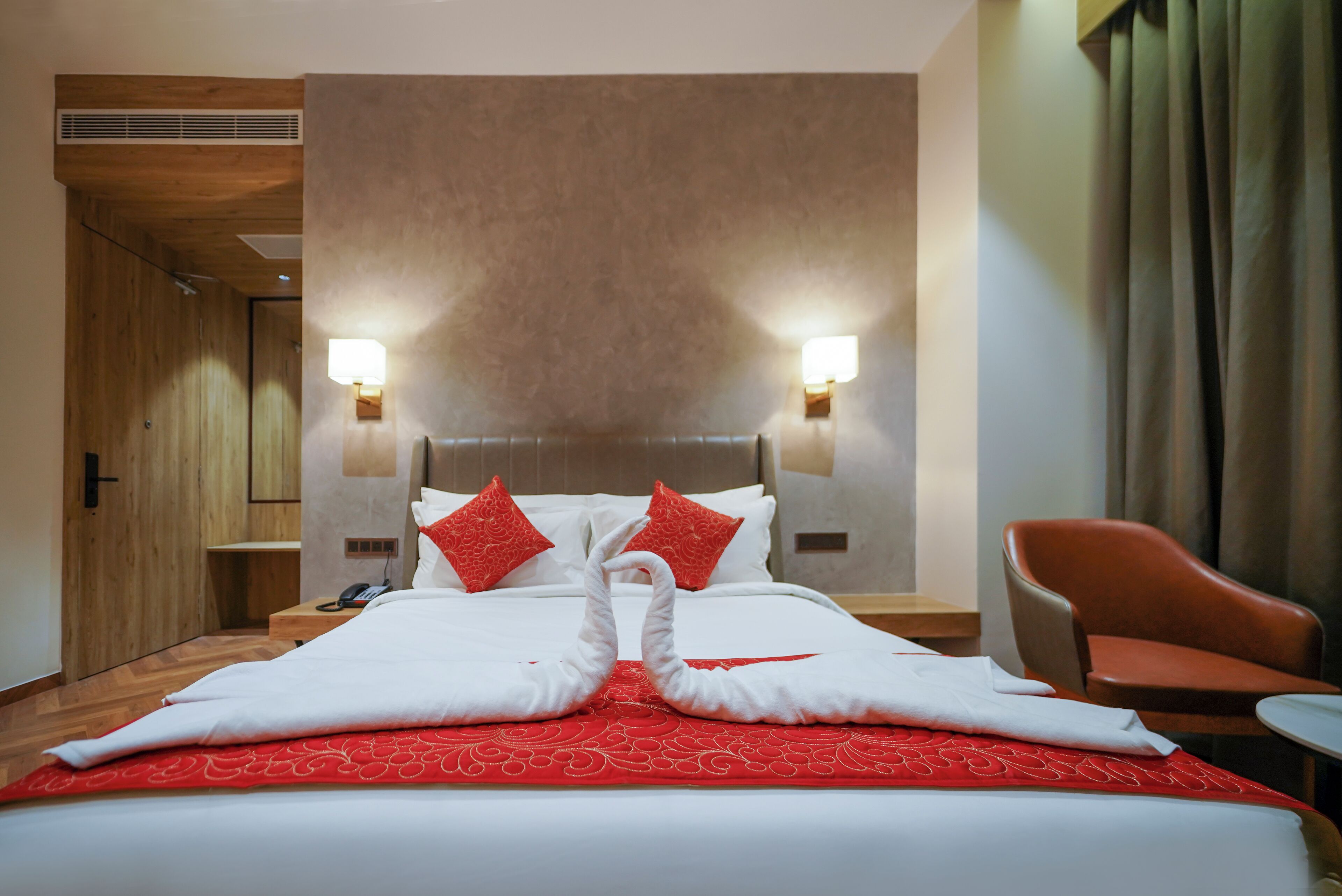 A hotel room with a large bed, two wall-mounted lamps, a wooden door, and a red chair.