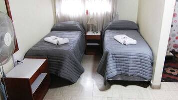 Una habitación con dos camas, una mesita de noche, una lámpara, un ventilador y una ventana con cortinas.