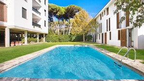 Una piscina rectangular con una escalera metálica, rodeada por un césped bien cuidado y edificios blancos con balcones.