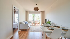 A modern living room with a sofa, dining table, and chairs. There is a balcony with a view of palm trees. - Costa Brava E-4 in Calella de Palafrugell (Palafrugell)
