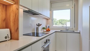 A modern kitchen with white cabinets, a wooden accent wall, a built-in oven, and a window with a view of greenery. - Costa Brava E-4 in Calella de Palafrugell (Palafrugell)
