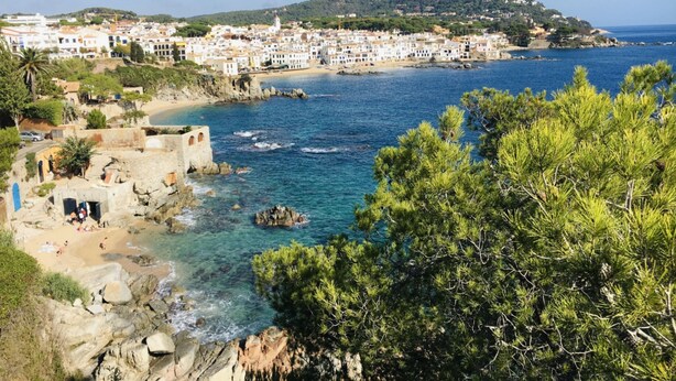 A coastal town with buildings along the shoreline, clear blue waters, and rocky terrain. - Costa Brava E-4 in Calella de Palafrugell (Palafrugell)