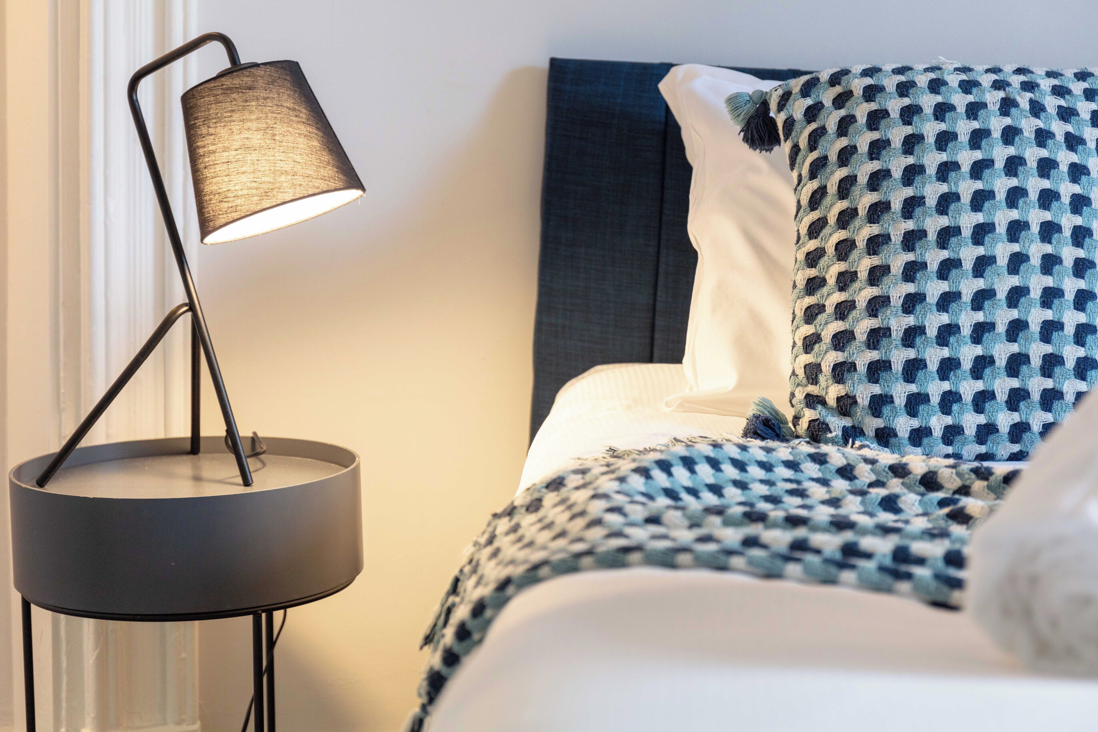 A neatly made bed with a patterned blanket and pillows, a modern floor lamp, and a dark headboard.
