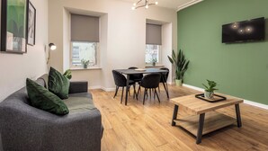 A modern living room with a grey sofa, a wooden coffee table, a dining table with chairs, a TV mounted on the wall, and a green accent wall. - The Annick Suite No.1 (Irvine)