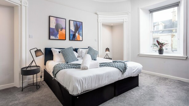 A modern bedroom with a large bed, two bedside tables, a window with a view, and abstract art on the wall. - The Annick Suite No.1 (Irvine)