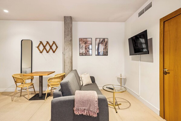 Una sala de estar moderna con un sofá gris, una mesa de comedor de madera con sillas de mimbre, un televisor montado en la pared y un pilar de concreto.