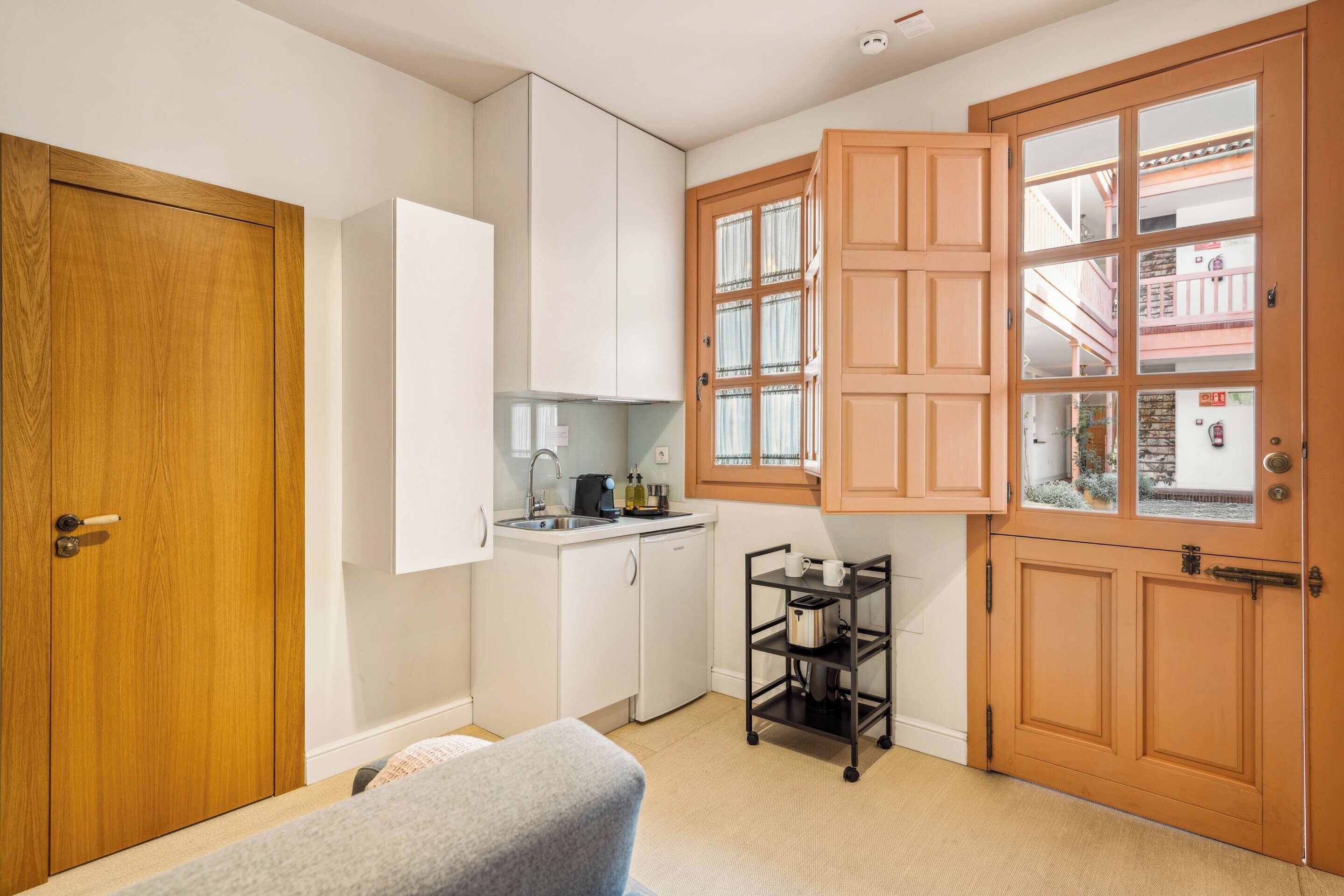 A compact kitchen with white cabinets, a sink, and a small countertop. There's a wooden door with a glass panel, and a metal cart with kitchen items.