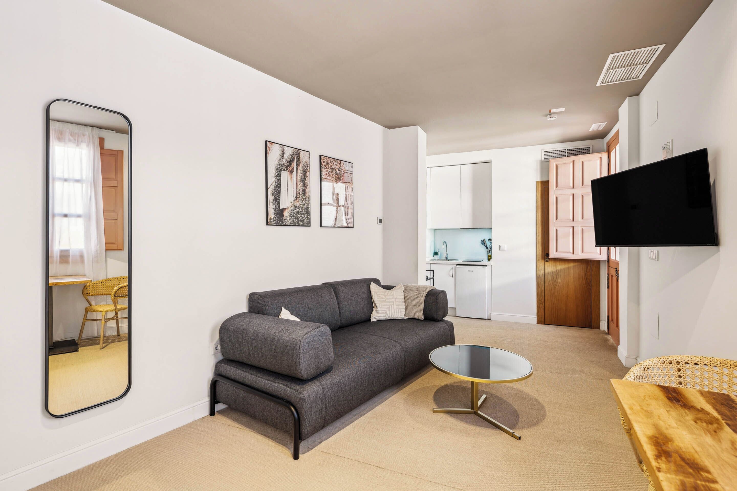 A modern living room with a grey sofa, a round coffee table, and a large mirror. There is a kitchen area visible in the background with white cabinets and a flat-screen TV mounted on the wall.