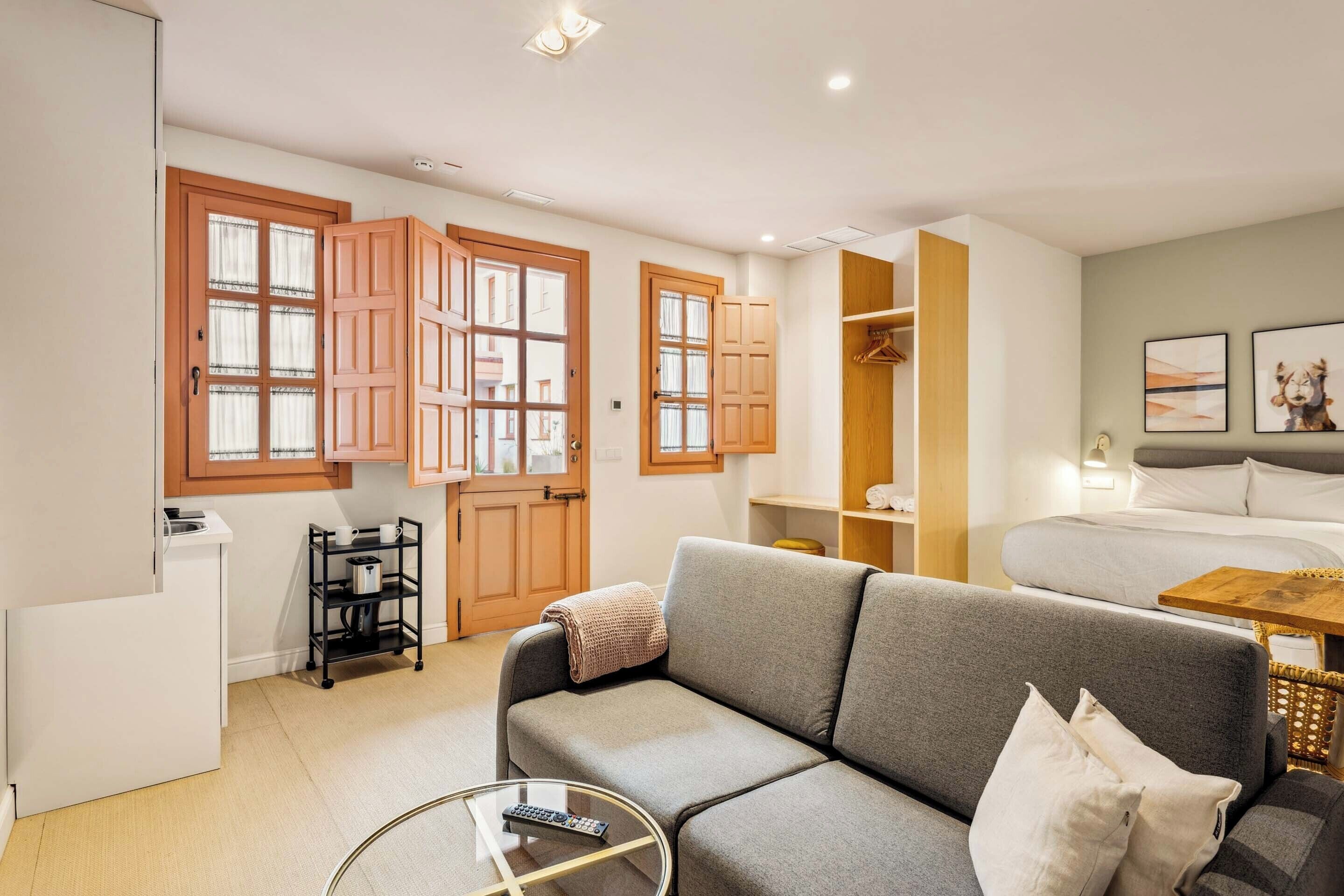 Una habitación de hotel moderna con un sofá gris, una mesita redonda, una cama con cabecero gris, puertas de madera y una pequeña kitchenette.