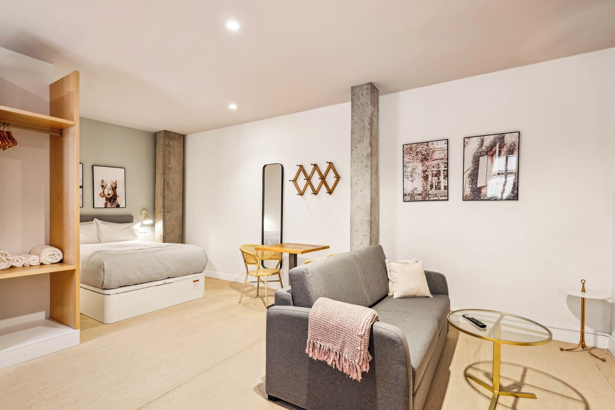 A modern room with a grey sofa, a bed with a wooden headboard, a small round table, and a wall-mounted mirror.