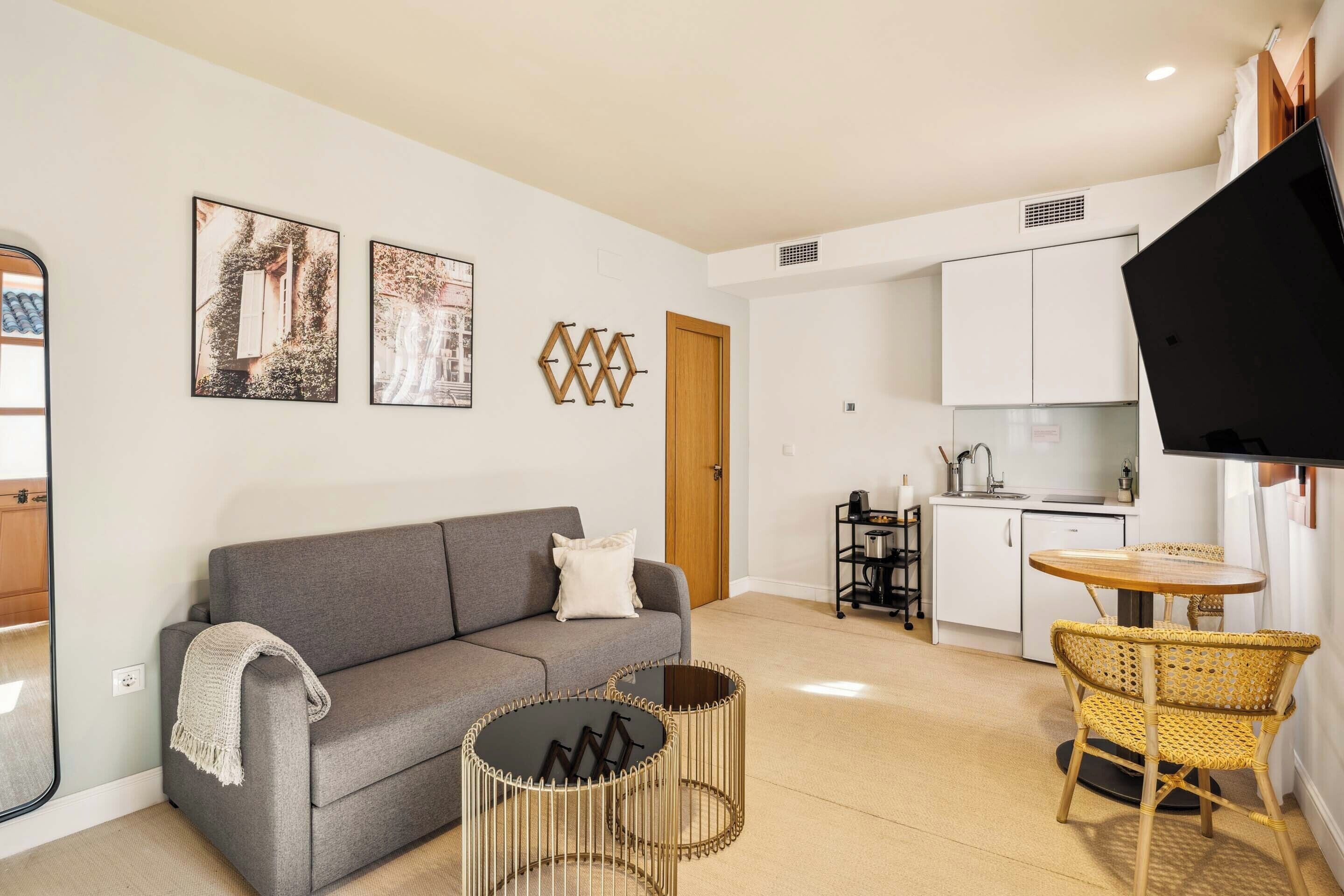 A modern living room with a grey sofa, a small round table, and a wicker chair. There is a kitchen area with white cabinets and a black TV mounted on the wall.