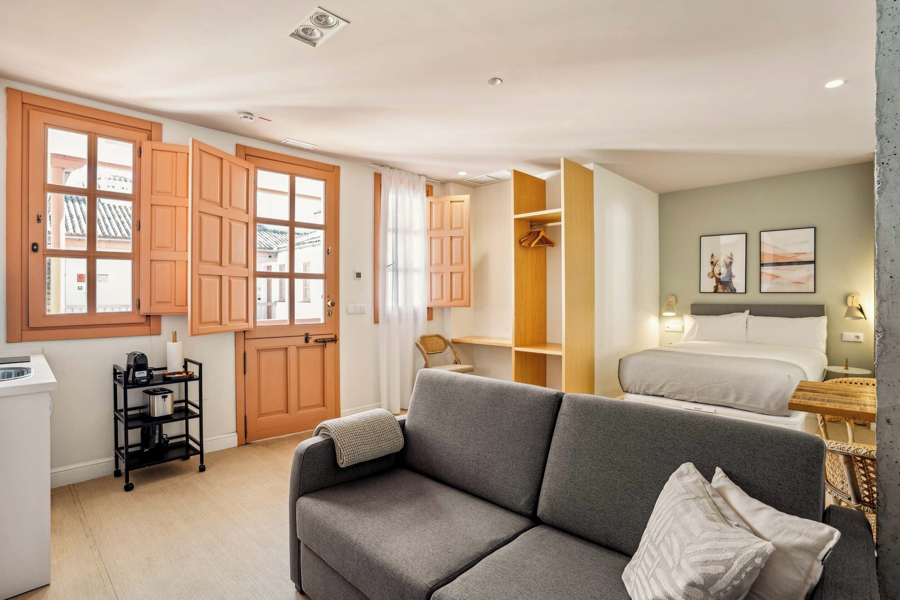 A compact hotel room with a grey sofa, a bed, a wooden wardrobe, and a kitchenette area.