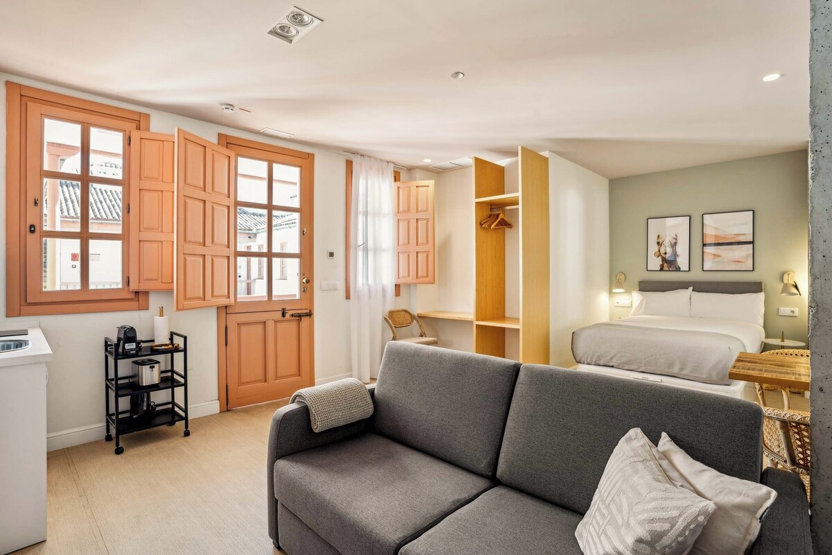 A compact hotel room with a grey sofa, a bed, a wooden wardrobe, and a kitchenette area.