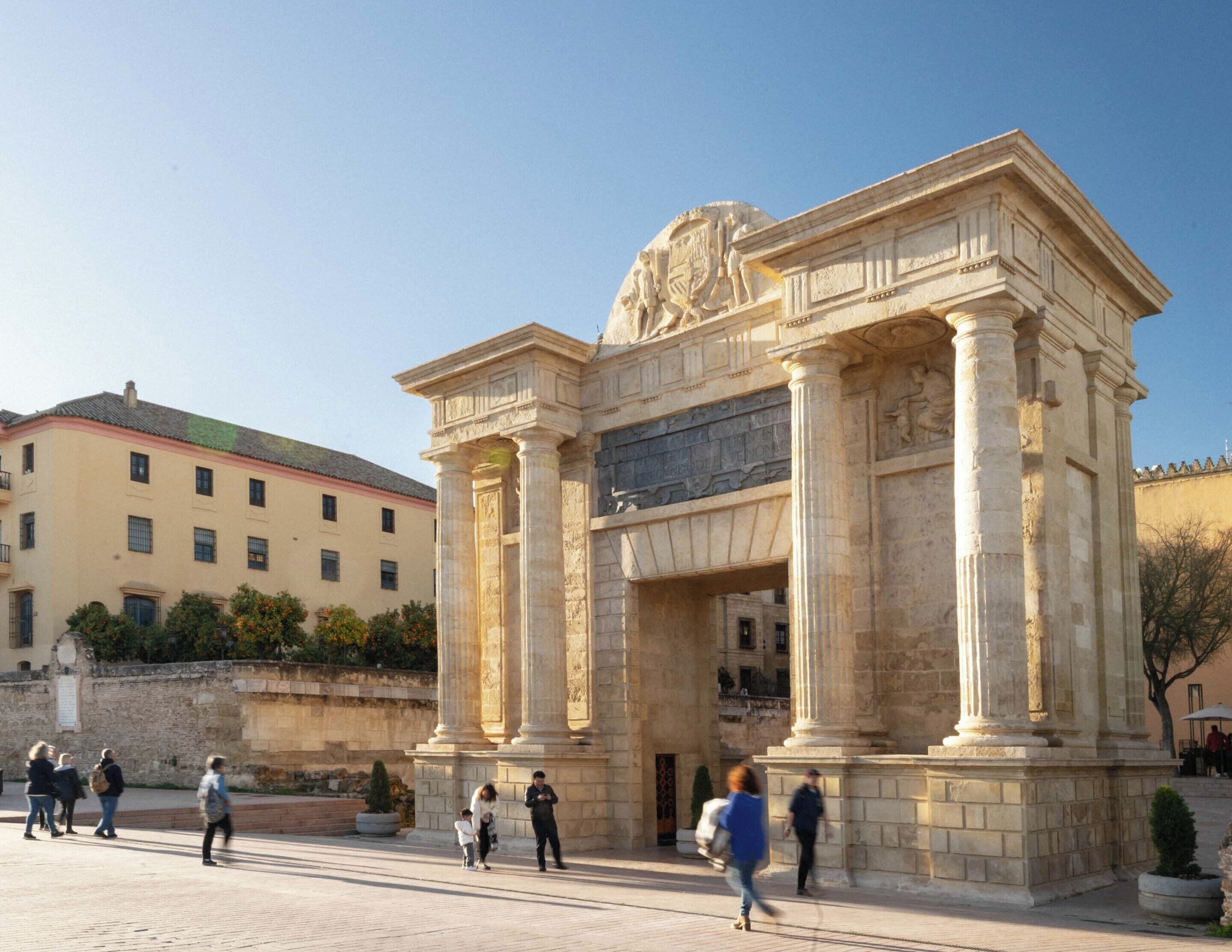 Un edificio clásico con columnas y un arco central, rodeado de una zona pavimentada con gente caminando.