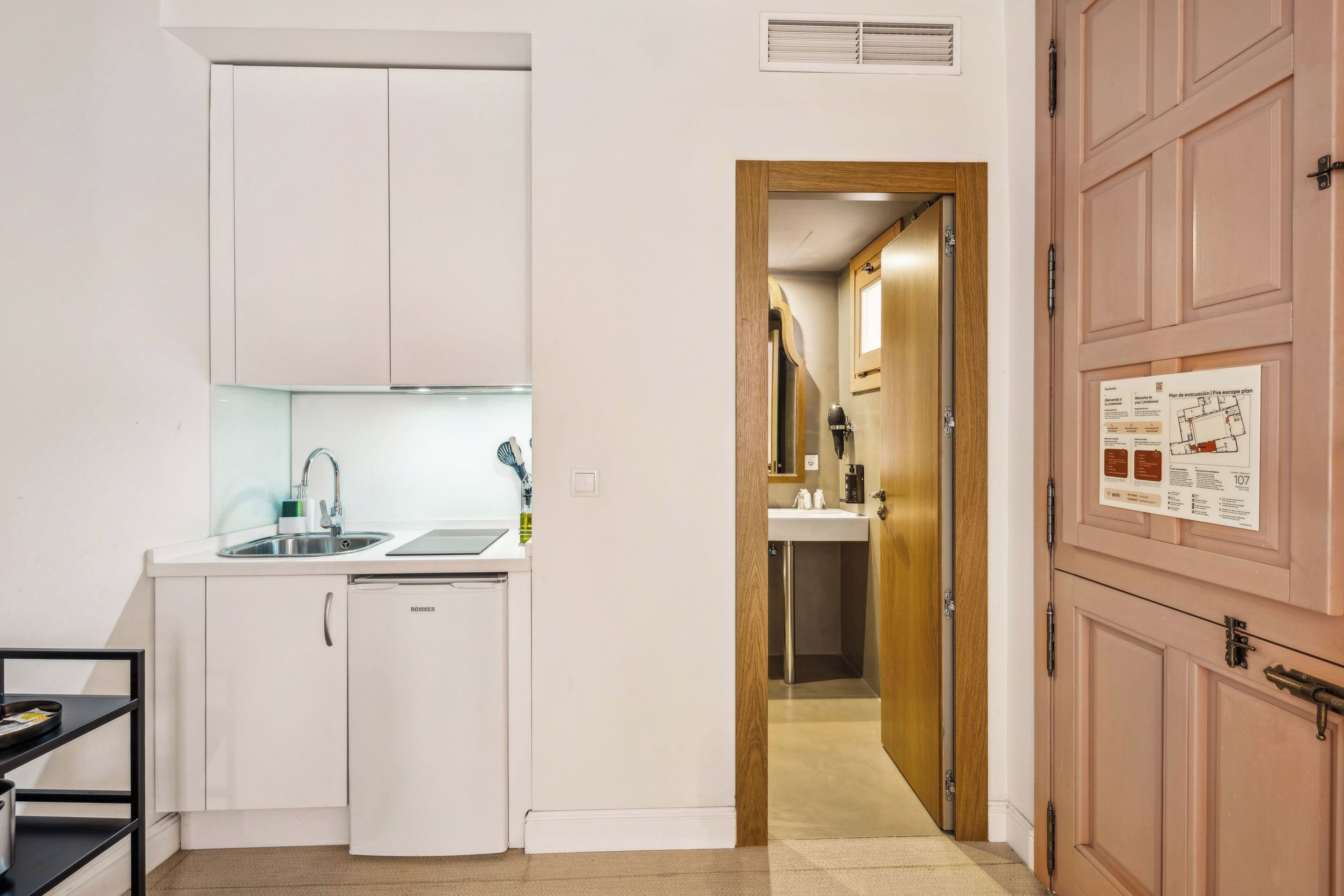 Cocina pequeña con armarios blancos, un lavabo y lavavajillas. Una puerta de madera da paso a un baño con lavabo y espejo.