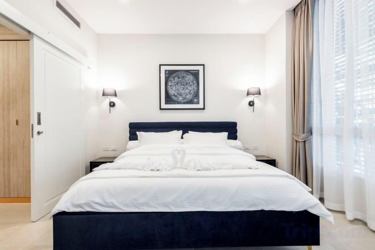 A bedroom with a bed, two bedside lamps, a framed artwork above the bed, and a window with curtains.