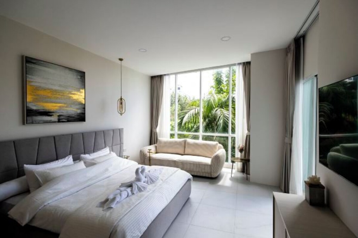 A modern bedroom with a large bed, a sofa, a painting, and a window with a view of greenery.