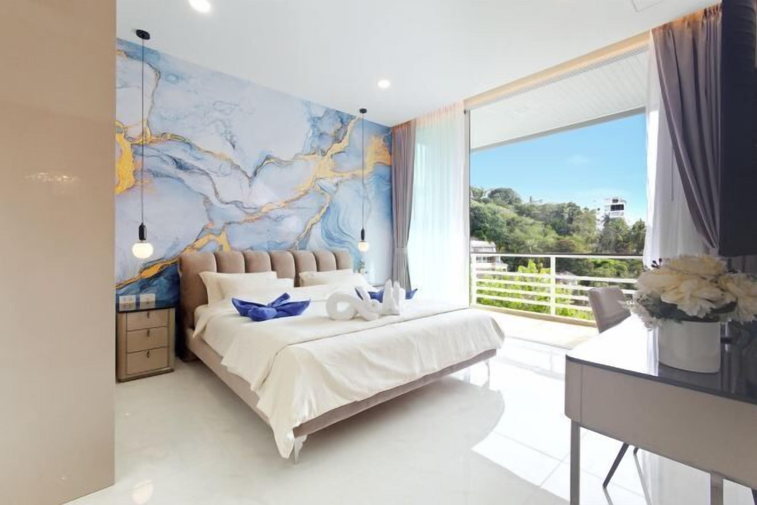 A modern bedroom with a large bed, a nightstand, a desk with a chair, and a view of greenery outside.