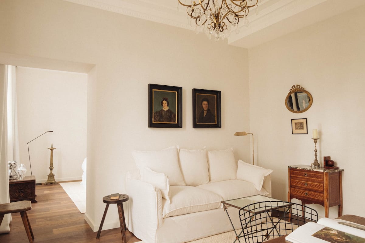 Een kamer met een witte bank, een houten bijzettafeltje, een klein rond tafeltje, een stoel, een vloerlamp en een kroonluchter.