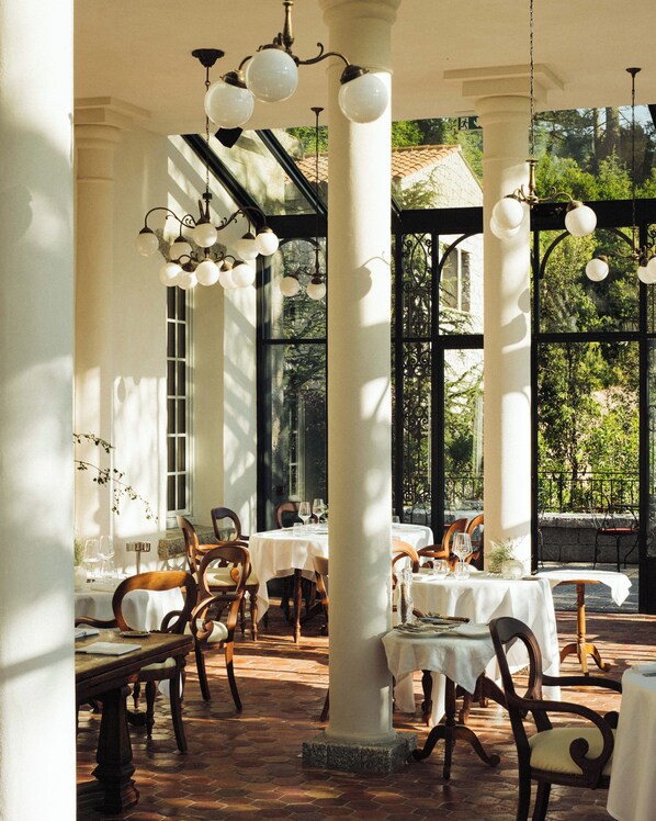 An indoor dining area with tables set for two, white columns, and hanging light fixtures. - Domaine Le Mouflon D'Or (Zonza)