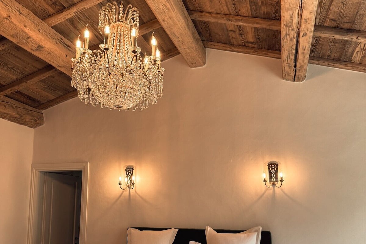 Een slaapkamer met een houten plafond, een kroonluchter, twee wandlampen, een bed met witte lakens en een nachtkastje.