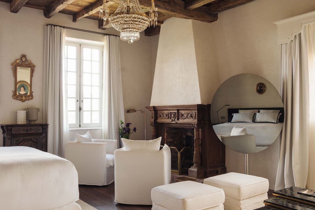 Een kamer met een bed, twee witte fauteuils, een ronde spiegel, een open haard en een kroonluchter.