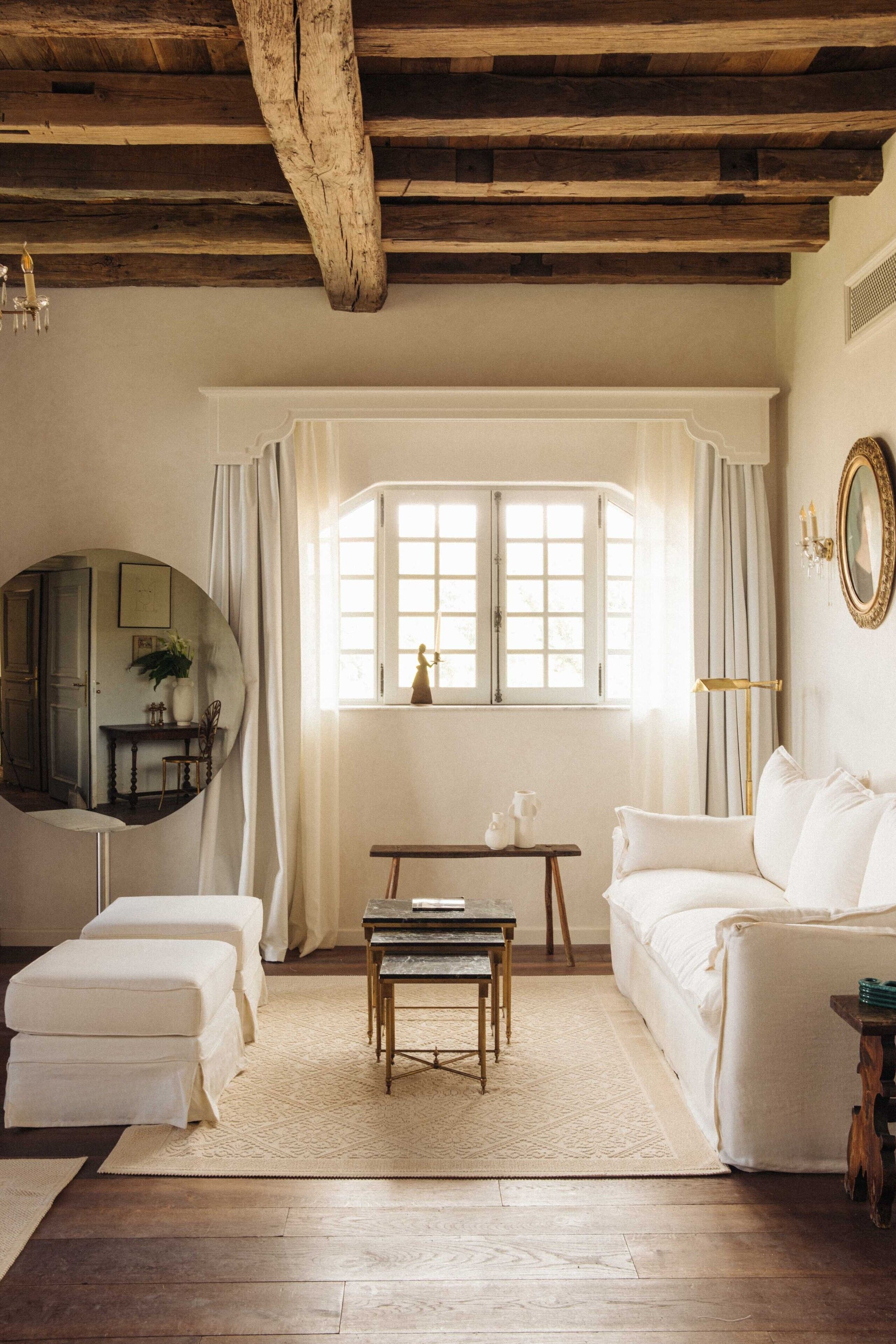 Una habitación con techo de madera, un sofá blanco, un espejo redondo, una ventana con cortinas y una mesita con un jarrón.