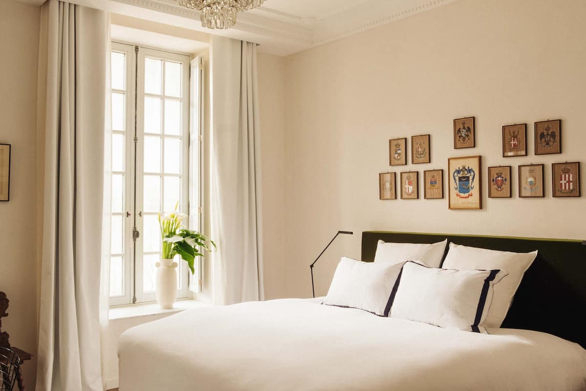 Een groot bed met witte lakens, een vaas met bloemen en een raam met gordijnen.