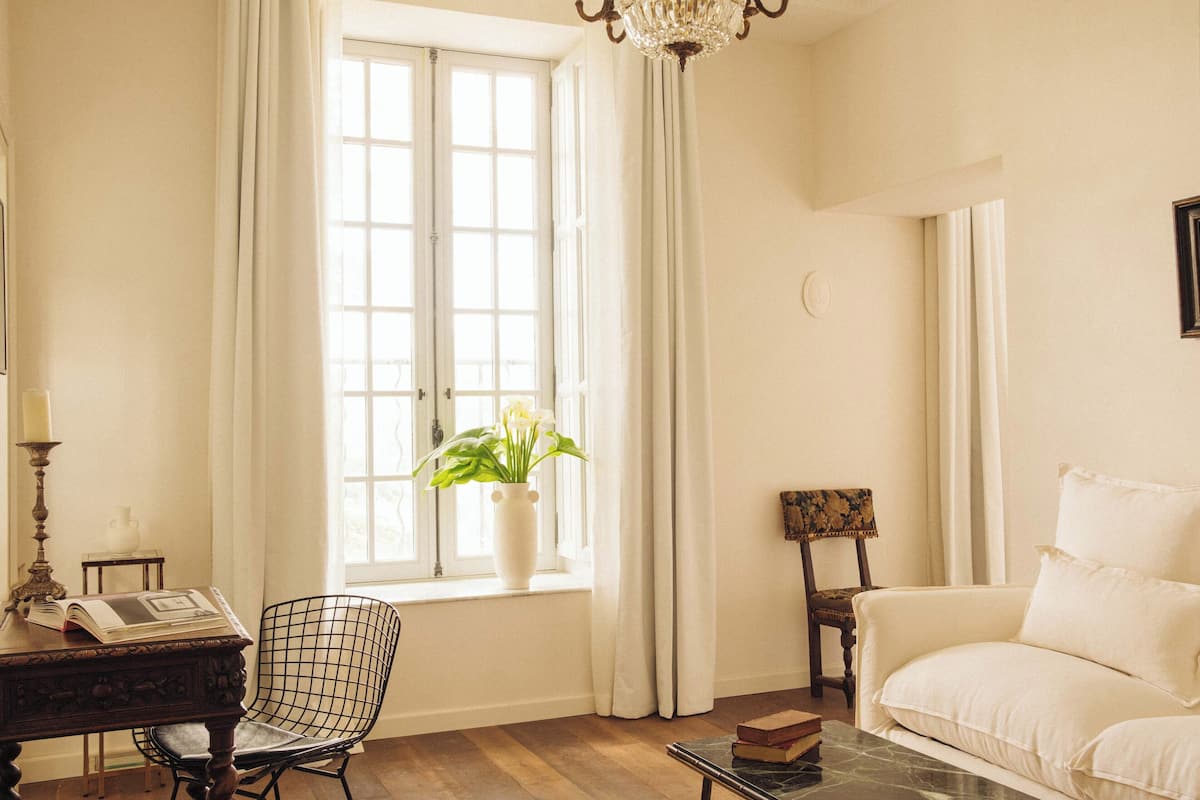 Een kamer met een groot raam, een witte bank, een houten stoel, een kleine tafel met boeken en een vaas met bloemen.