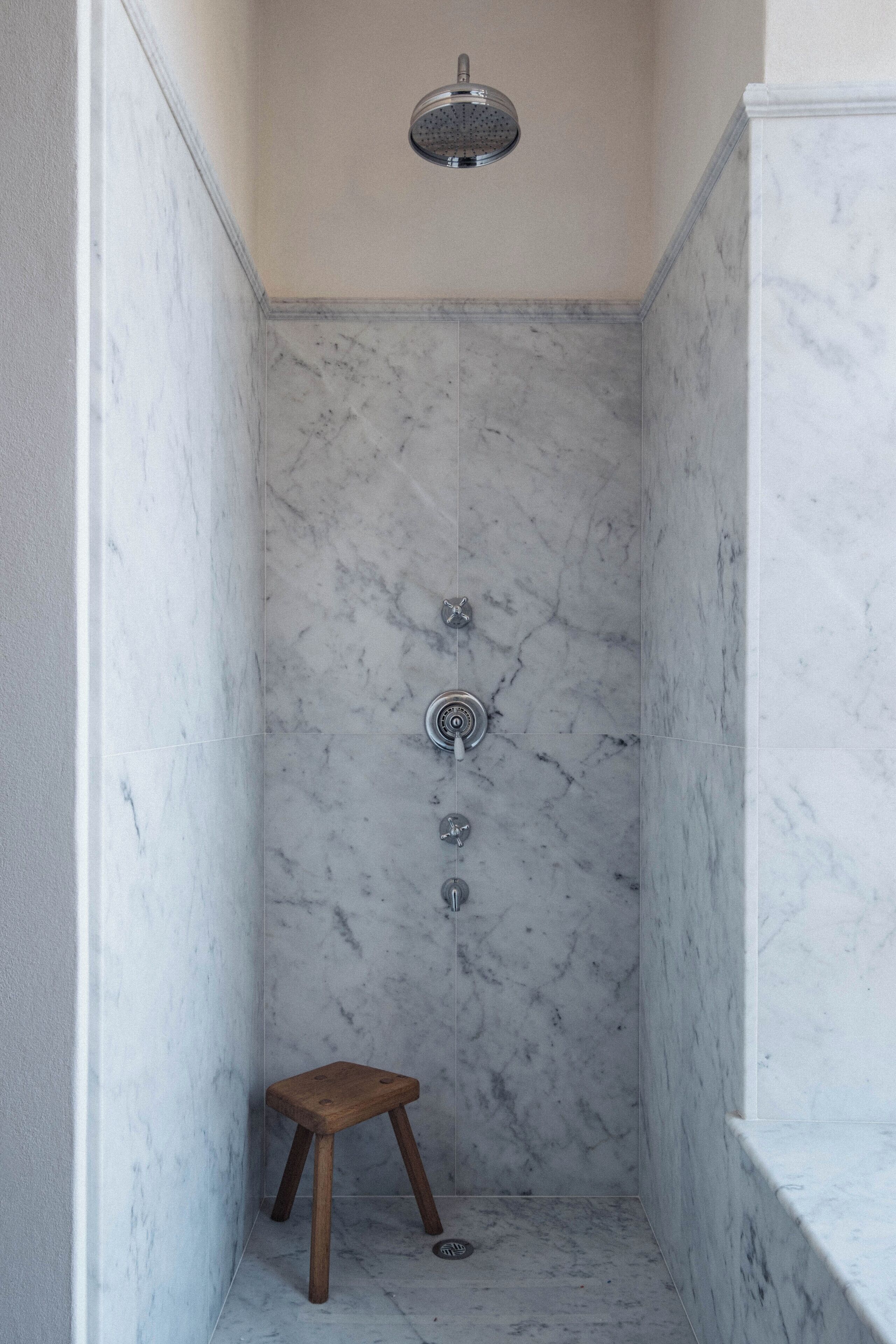 Un baño con ducha, paredes de mármol y un taburete de madera.